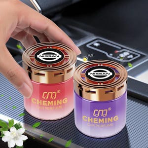 Könnte beinhalten: Zwei Auto-Lufterfrischer mit goldfarbenen Deckeln und dem Nissan-Logo. Einer ist rot, der andere lila, beide mit dem Text "CHEMING CAR PERFUME" in Gold. Die zylindrischen Erfrischer stehen auf einer dunklen Oberfläche.