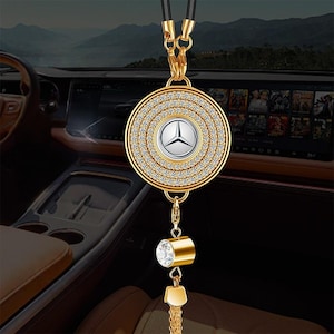 Puede incluir: Amuleto de coche dorado con un colgante circular. Presenta un logotipo plateado de Mercedes-Benz en el centro, rodeado de cristales transparentes. El amuleto cuelga de un cordón negro e incluye un cristal y una borla colgantes.
