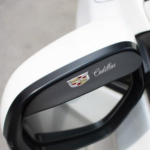 Könnte beinhalten: Weißer Cadillac-Seitenspiegel mit schwarzem Zierrat. Das Cadillac-Emblem befindet sich auf einer schwarzen Platte, mit dem Markennamen in silberner Schrift. Das Emblem zeigt ein Wappen-Design mit goldenen, roten und schwarzen Akzenten.