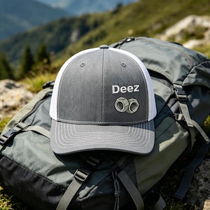 May include: A grey and white trucker hat featuring the word "Deez" and a graphic of two nuts. The hat is on a backpack with a mountain backdrop. The hat has a mesh back and a curved peak.