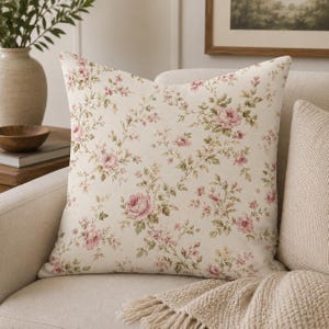 Può includere: Cuscino quadrato con sfondo crema e motivo floreale di rose rosa e foglie verdi. Il cuscino è su un divano color crema, con una coperta beige in vista.