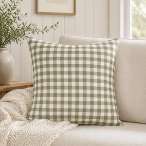 May include: Square throw pillow with a green and white gingham pattern. The pillow is on a beige sofa with a cream-colored knitted throw. A vase with greenery is visible in the background.