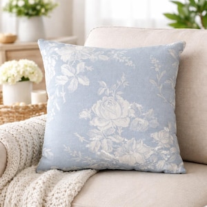 Puede incluir: Un cojín cuadrado azul claro con un estampado floral blanco con rosas y otras flores. El cojín está colocado sobre un sofá beige, con una manta blanca de punto visible.