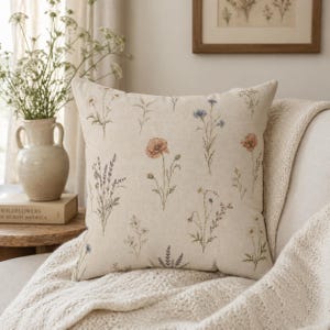 May include: Square cushion with a beige background and a floral pattern featuring wildflowers in shades of orange, blue, and purple. The cushion is placed on a white knitted blanket.