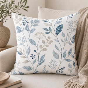 May include: Square throw pillow with a white background and a botanical pattern featuring blue and tan leaves and berries. The pillow is placed on a neutral-colored sofa, with a beige throw blanket visible in the background.