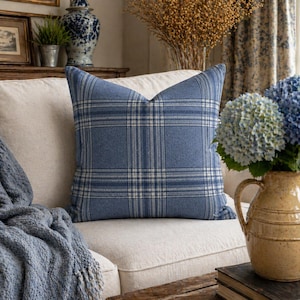 May include: A blue plaid throw pillow with a white and blue grid pattern rests on a cream-colored sofa. A blue knitted throw blanket and a vase of blue hydrangeas are also visible.