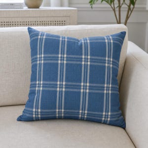Op de afbeelding: Een vierkant blauw sierkussen met een ruitpatroon in wit en lichtbeige. Het kussen ligt op een lichtbeige bank. Het kussen heeft een zacht, getextureerd uiterlijk.