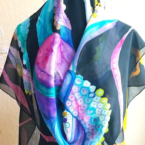 May include: A black silk scarf with a colorful octopus design. The octopus is painted in shades of blue, pink, green, and yellow. The scarf is a square shape and has a fringed edge.