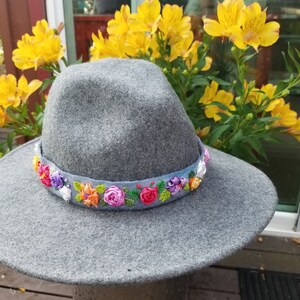 Może przedstawiać: Szary filcowy kapelusz fedora z szerokim rondem i kolorową wstążką z kwiatami wokół korony.