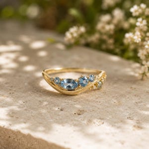 May include: A gold ring with a wave design, set with multiple blue gemstones. The ring features an oval-shaped center stone and smaller round stones, creating a flowing, elegant look. The ring is displayed on a light-colored surface.