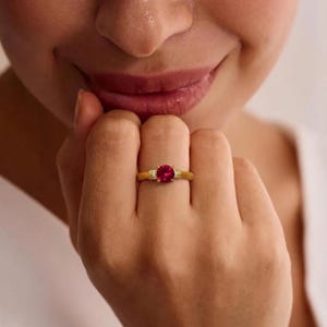 May include: A gold ring with a large, round, red gemstone as the centerpiece. The ring has small, clear accent stones on either side of the main stone. The ring is on a person's finger.