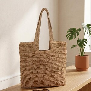 May include: A woven tote bag in a natural tan color. The bag has a rectangular shape with a wide opening and two long handles. Sunglasses and a notebook rest on a wooden bench in front of the bag. A potted plant is in the background.