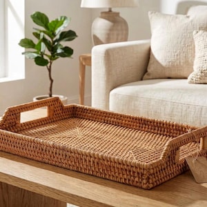 May include: A rectangular woven rattan tray with integrated handles. The tray is light brown and sits on a wooden table. A blank tag hangs from one of the handles. The tray is a decorative home decor item.