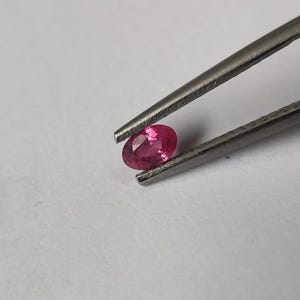 以下が含まれることがあります： 銀色の金属製ピンセットで保持された、小さな楕円形の鮮やかなピンク色の宝石。宝石には光を反射する複数のファセットがあり、無地の明るい背景に対して明るく半透明な外観を作り出しています。