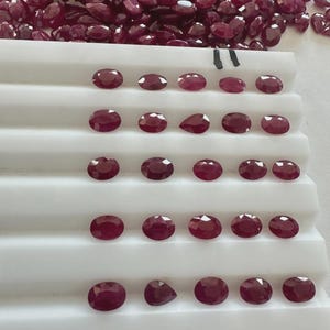 Puede incluir: Una colección de rubíes facetados de forma ovalada, de varios tamaños y cortes. Las piedras preciosas son de un color rojo intenso y rico, exhibidas sobre una superficie blanca escalonada. Una pila de rubíes sueltos se encuentra encima.