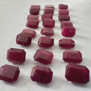 May include: A collection of faceted, rectangular-cut gemstones in varying shades of red. The gems are arranged on a white surface, showcasing their polished surfaces and internal clarity. The gemstones appear to be rubies.