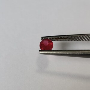 May include: A small, round, faceted gemstone with a vibrant red color held securely between the metal tips of a pair of tweezers. The stone exhibits a translucent quality with visible internal inclusions and a polished, reflective surface texture.