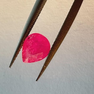 May include: A faceted, teardrop-shaped, bright red gemstone is held by a pair of tweezers. The gem has a polished surface and appears to be a ruby. The tweezers are dark metal and are holding the gem against a light background.