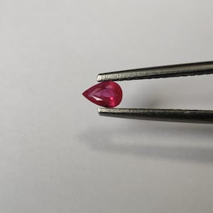 Op de afbeelding: Een peervormige, gefacetteerde edelsteen met een levendige, dieprode kleur, stevig vastgehouden tussen de twee metalen punten van een zilverkleurige pincet. De edelsteen heeft heldere, scherpe randen en een gepolijst oppervlak dat licht reflecteert.