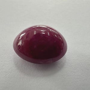 May include: A smooth, oval gemstone in a deep burgundy hue. The surface is reflective, showing light. The gem is set against a plain white background. This cabochon could be used in jewellery.