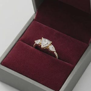 May include: A gold ring with three clear, triangular-cut stones, presented in a gray jewelry box with a burgundy velvet interior. The ring's band has a textured design. The box is open, showcasing the ring against the plush background.