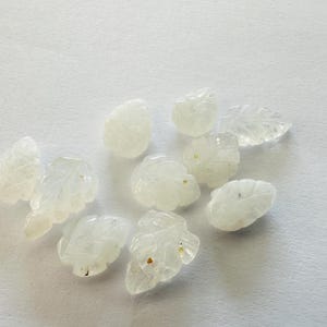 May include: A collection of small, translucent, leaf-shaped gemstones. The gemstones are carved with intricate details, showcasing the veins and contours of the leaves. The stones are scattered on a white surface, creating a delicate and natural aesthetic.