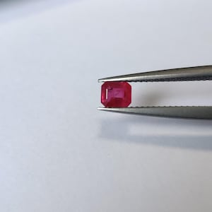 Puede incluir: Una gema de rubí rojo de corte rectangular sostenida firmemente por unas pinzas de metal. La piedra preciosa muestra un tono carmesí profundo y saturado con facetas internas visibles y una superficie pulida y reflectante.