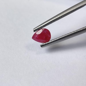 May include: A pear-shaped, faceted red gemstone held by a pair of metal tweezers. The stone exhibits a deep, vibrant crimson hue with multiple geometric facets that reflect light across its surface, showcasing a clear and polished appearance.