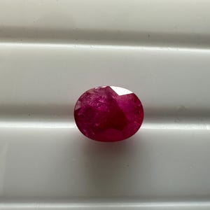 May include: A faceted, oval-shaped ruby gemstone. The ruby is a deep, saturated red color, with visible internal inclusions. The gemstone is set against a white background, highlighting its color and clarity. This gemstone is suitable for jewelry making.