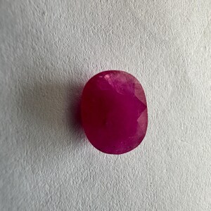 May include: A faceted, oval-shaped ruby gemstone. The gem is a deep, saturated red color, with a polished surface that reflects light. The gemstone is set against a textured, off-white background.
