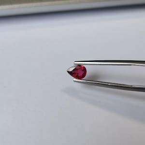 Puede incluir: Una piedra preciosa roja tallada en forma de pera, sostenida por unas pinzas de metal plateado. La piedra muestra un tono carmesí vibrante y translúcido con múltiples facetas que reflejan la luz, exhibiendo una superficie clara y pulida.