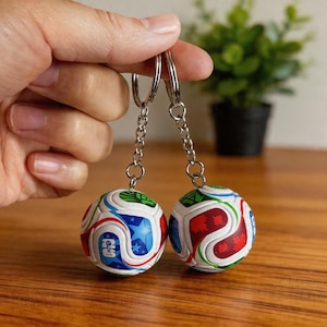 Puede incluir: Dos llaveros de balón de fútbol. Cada llavero presenta un balón de fútbol en miniatura con una base blanca y diseños coloridos, que incluyen estrellas azules, remolinos rojos y detalles verdes. Los llaveros están sujetos por un anillo y una cadena plateados.