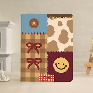 May include: A rectangular notebook featuring a patchwork design with a blue section containing a brown button, a brown plaid section with two red bows, a beige and brown cow print section, and a dark red section with a yellow smiley face.
