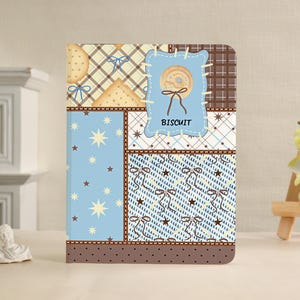 Pode incluir: Um caderno retangular com design de patchwork em tons de azul claro, castanho e creme. A capa apresenta padrões de estrelas, laços e xadrez, com um emblema azul central com a palavra BISCUIT e uma ilustração de uma bolacha redonda.