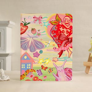 May include: A rectangular notebook featuring a colorful illustration of a large red strawberry, a purple moth, a pink snail, a small house, butterflies, stars, and a bow against a pastel yellow and green background with various small floral motifs.