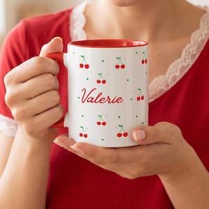 Puede incluir: Taza de cerámica blanca con interior y asa rojos, decorada con cerezas rojas y lunares verdes. El nombre "Valerie" está escrito en rojo en la parte delantera. La taza la sostiene una persona que lleva una camiseta roja.