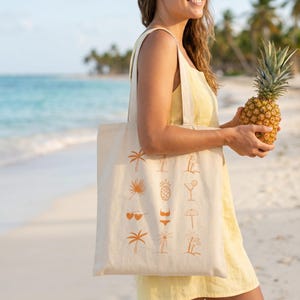 Puede incluir: Un bolso tote beige con ilustraciones tropicales naranjas, incluyendo palmeras y una piña. Una piña entera se sostiene en la mano derecha. El fondo muestra una playa y el océano.