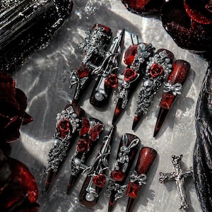 Peut inclure: Une collection de faux ongles rouge foncé et noirs avec des ornements argentés complexes. Les ongles présentent des motifs tels que des crânes, des roses et des nœuds, avec une finition pailletée. Le texte "Meltynails" est visible en haut à droite.
