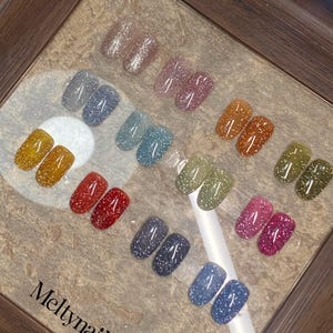 May include: A display of various glitter nail polish colors, including silver, pink, orange, green, and blue. The nail polish is arranged in pairs, showcasing a range of shades and finishes. The brand name "Meltnails" is visible.