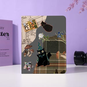 Pode incluir: Um caderno retangular com design dividido, apresentando uma ilustração bege de um cômodo e um padrão xadrez verde-escuro. Exibe um gato preto com um chapéu de mago estrelado, um segundo gato preto, cogumelos, flores, um pingente em forma de estrela e uma placa com a palavra PEACE.