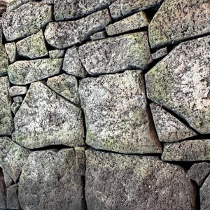 Pu&ograve; includere: Un muro in pietra costruito con blocchi di granito di forma irregolare e grezza in tonalit&agrave; grigio e antracite. Le pietre presentano una consistenza maculata e usurata con sottili chiazze di muschio verde nelle fessure profonde.
