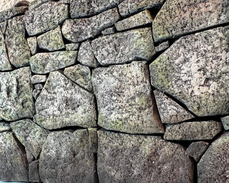 Pu&ograve; includere: Un muro in pietra costruito con blocchi di granito di forma irregolare e grezza in tonalit&agrave; grigio e antracite. Le pietre presentano una consistenza maculata e usurata con sottili chiazze di muschio verde nelle fessure profonde.