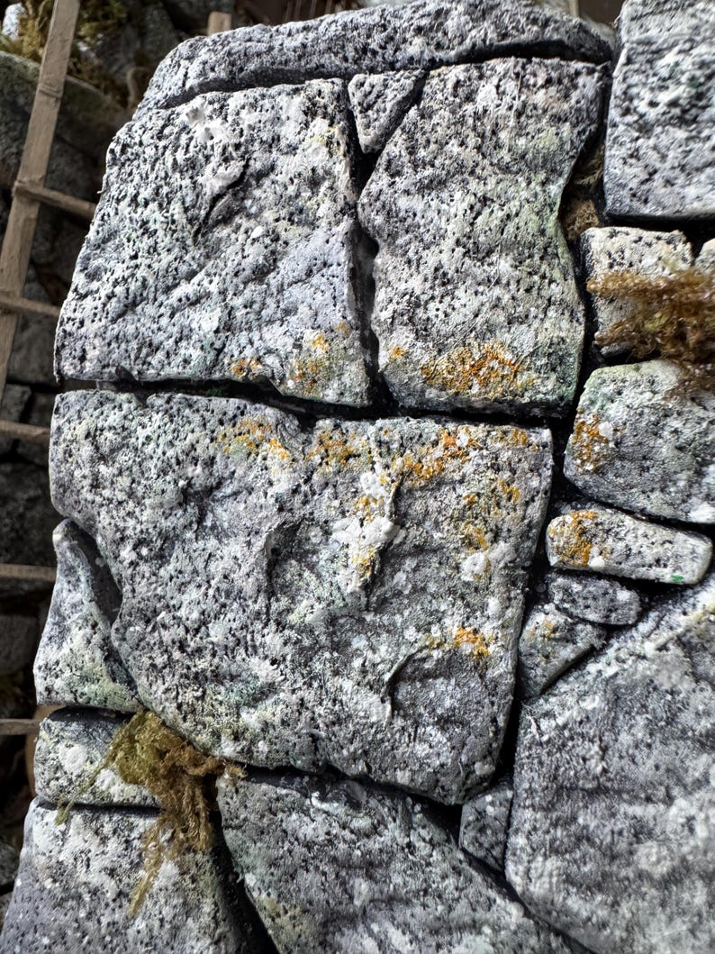 Pu&ograve; includere: Un muro in pietra costruito con blocchi di granito grigio irregolari e ruvidi. La superficie delle pietre presenta un motivo punteggiato con macchie di licheni gialli e verdi. Le pietre sono assemblate con spazi visibili tra loro.