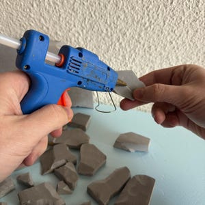 Pu&ograve; includere: Una pistola per colla a caldo blu con grilletto arancione e stick di colla trasparente inserito, che applica adesivo su un piccolo frammento di pietra grigia irregolare. Altri pezzi di pietra grigia simili sono sparsi sulla superficie.