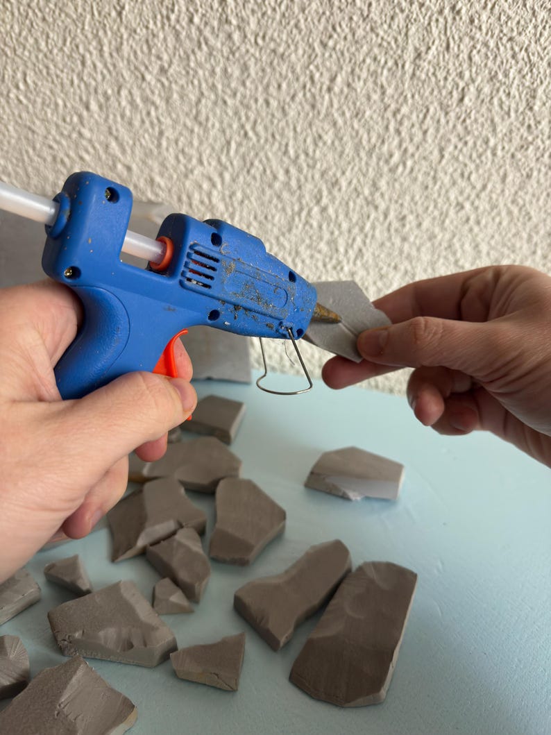 Pu&ograve; includere: Una pistola per colla a caldo blu con grilletto arancione e stick di colla trasparente inserito, che applica adesivo su un piccolo frammento di pietra grigia irregolare. Altri pezzi di pietra grigia simili sono sparsi sulla superficie.