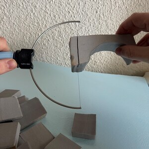 Pu&ograve; includere: Un taglia-schiuma a filo manuale con manico azzurro e un connettore nero con l'etichetta HW-11071, che taglia un pezzo curvo di schiuma grigia. Diversi piccoli blocchi quadrati di schiuma grigia sono sparsi sotto l'utensile.