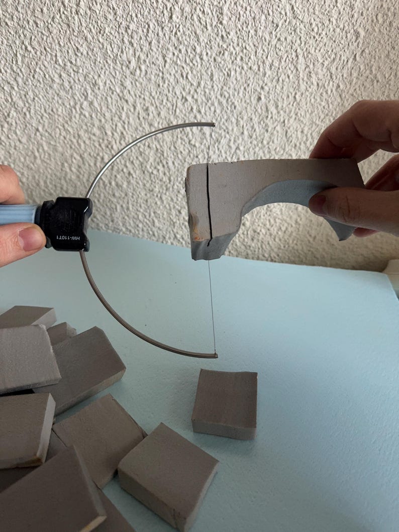 Pu&ograve; includere: Un taglia-schiuma a filo manuale con manico azzurro e un connettore nero con l'etichetta HW-11071, che taglia un pezzo curvo di schiuma grigia. Diversi piccoli blocchi quadrati di schiuma grigia sono sparsi sotto l'utensile.