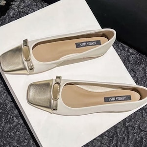 Peut inclure: Une paire de ballerines blanches avec des bouts carrés dorés et une boucle décorative. Les chaussures ont une semelle intérieure de couleur neutre avec le texte "AINDA MISS". Les ballerines sont présentées sur une surface blanche.