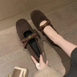 May include: A pair of dark brown textured fabric flats featuring a rounded toe, a strap across the instep with a small gold-toned rose-shaped embellishment, and a black interior lining. The shoes have a soft, fuzzy exterior finish.