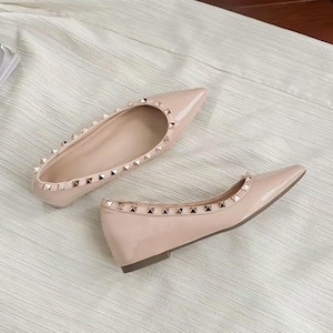 May include: A pair of beige pointed-toe flats featuring a glossy finish and a row of small, metallic pyramid-shaped studs along the top edge of the opening. The shoes have a low, flat heel and a sleek, streamlined silhouette.